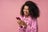 Beautiful smiling African American woman using smartphone playing mobile game isolated on pink background. Happy stylish female holding mobile phone shopping online with sale, copy space Sending gift card via text