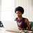Woman of color sitting on a wooden table typing on a silver laptop.