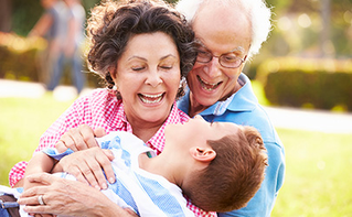 Smiling grandparents joyfully hugging their young grandson while sitting on the grass in a sunny park.