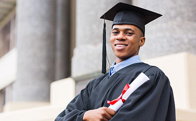 Smiling graduate wearing a cap and gown, holding a rolled-up diploma tied with a red ribbon, standing in front of a building with large columns.