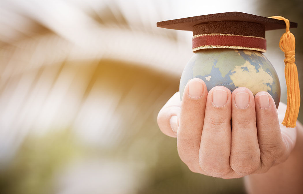Hand holding a small globe with a graduation cap on top, against a blurred background with light and shadow patterns.